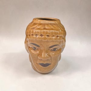 May include: A tan ceramic vase shaped like a human head with a textured band across the forehead. The face features dark painted eyes and dark lips, with a smooth, glossy finish covering the entire surface of the vessel, including the opening.
