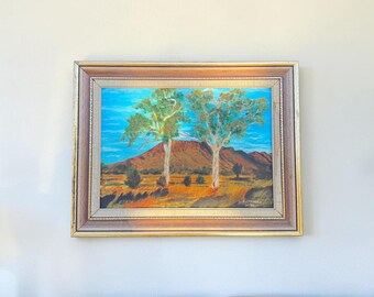 May include: An oil painting of a landscape featuring two trees in the foreground with a mountain in the background. The painting has a gold and brown frame. The sky is blue, and the trees have green foliage.