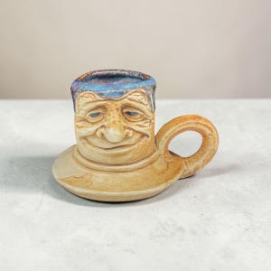 May include: A ceramic mug featuring a sculpted human face with a smiling expression. The mug has a tan, textured finish and a wide, flat base. The rim is glazed in a mottled blue and purple tone, and a curved handle extends from the right side.