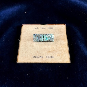 May include: A rectangular brooch featuring an iridescent N.Z. Paua shell inlay set in sterling silver. The piece is mounted on a square beige display card with the printed text N.Z. PAUA SHELL at the top and STERLING SILVER at the bottom.