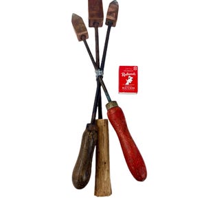 May include: A set of vintage metal tools with wooden handles, including a red-handled tool and a matchbox. The tools have different shaped metal heads and are bound with wire. The matchbox is red with white text.