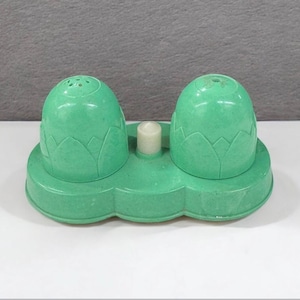 May include: A vintage green salt and pepper shaker set. The set features two egg-shaped shakers with small holes on top, on a matching base. A small, white candle-shaped item is between the shakers.