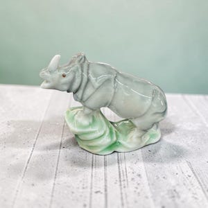 May include: A small ceramic rhinoceros figurine with a glossy finish. The rhinoceros is light grey with subtle green undertones and is depicted standing on a textured, light green base. The figure features a single horn and a small brown eye.