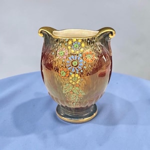 May include: A small, decorative vase with a flared top and a pedestal base. The vase is primarily brown with a floral design in shades of yellow, blue, green, and orange. The rim and base are trimmed with gold accents. The vase is approximately 13 cm tall.