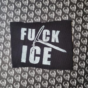 Puede incluir: Parche rectangular negro con las palabras "FUCK ICE" en blanco y un gráfico de un piolet. El parche está sobre un fondo de calaveras.