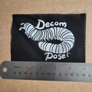 May include: Black fabric patch with the words "Decom Poser" above and below a white centipede illustration. The patch is approximately 4 inches wide.