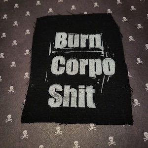 May include: Black fabric patch with the words "Burn Corpo Shit" in distressed white lettering. The patch has a raw, unfinished edge. The background is a dark gray pattern of white skulls and crossbones.