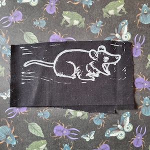 May include: A black fabric patch with a white rat illustration. The rat is depicted with an open mouth. The patch is rectangular with unfinished edges. The background is a dark grey fabric with insect and frog patterns.