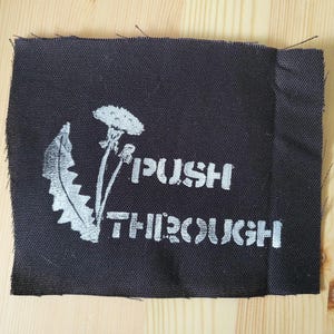 Puede incluir: Un parche de tela negra con texto blanco y una ilustración de diente de león. Las palabras "PUSH THROUGH" están impresas en una fuente en negrita sin serifa. El diente de león está a la izquierda.