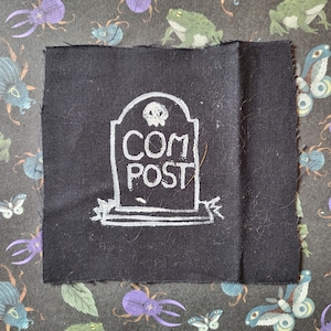 Puede incluir: Un parche de tela negro con un contorno blanco de una lápida que dice "COMPOST".