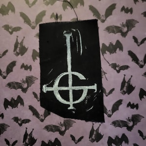 May include: A black fabric patch featuring a white inverted pentagram with a cross inside. The patch is displayed against a purple background with a pattern of bats.