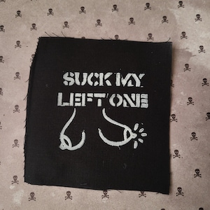 Puede incluir: Parche cuadrado negro con texto blanco que dice "SUCK MY LEFT ONE" y un gráfico de un pezón y areola estilizados. El parche está sobre un fondo con un patrón repetido de calaveras.