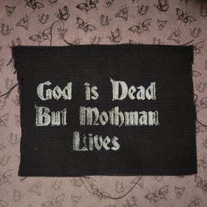 Op de afbeelding: Zwarte stoffen patch met witte tekst die "God is dood, maar Mothman leeft" zegt.