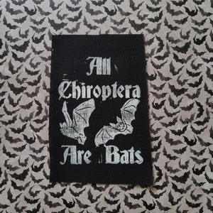 May include: Black rectangular patch with the text "All Chiroptera Are Bats" in a gothic font. The patch features two bat illustrations in white. The background is a pattern of gray bats.
