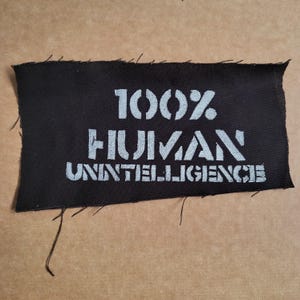 以下が含まれることがあります： 黒い長方形のパッチ。白いステンシル風の文字で「100% HUMAN UNINTELLIGENCE」と書かれています。パッチは未加工の端があり、ベージュ色の背景に配置されています。