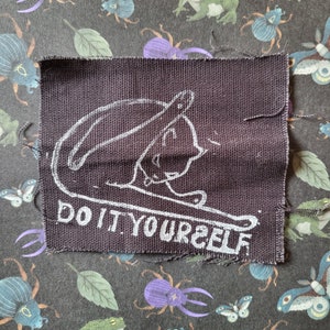 May include: A black fabric patch with white embroidery of a cat curled up and the text "DO IT YOURSELF".