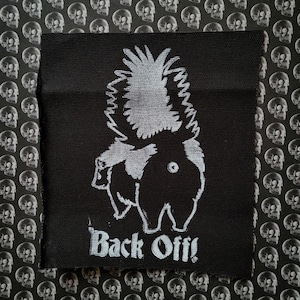 May include: Black and white iron-on patch with a cartoon image of a creature with a large, spiky tail and the text "Back Off!"