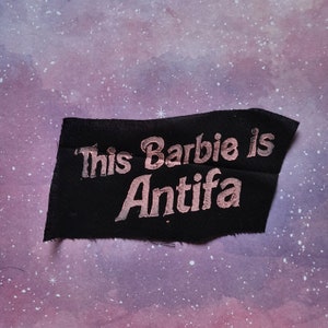 Pode incluir: Um patch de tecido preto com a frase "This Barbie is Antifa" impressa em ouro rosa metálico. O patch tem uma borda crua e está sobre um fundo roxo e branco.