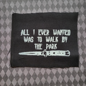 Puede incluir: Parche rectangular negro con el texto blanco "ALL I EVER WANTED WAS TO WALK BY THE PARK" y una ilustración blanca de un cuchillo. Sobre un fondo gris con patrón de diamantes.