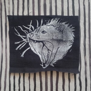 Op de afbeelding: Een zwarte stoffen patch met een witte afbeelding van een dier met een grote bek en scherpe tanden. De vacht van het dier is afgebeeld met een textuurpatroon.