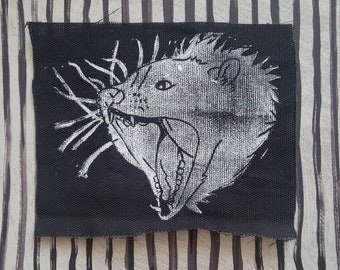 May include: A black fabric patch with a white image of an animal with a large mouth and sharp teeth. The animal's fur is depicted with a textured pattern.