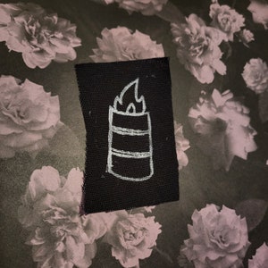 Peut inclure: Un écusson en tissu noir avec une illustration blanche dessinée à la main d'un baril avec des flammes. L'écusson est posé sur un fond de motif floral dans des tons de gris et de blanc.