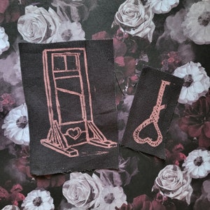 Puede incluir: Dos parches de tela negra con diseños rosas. Un parche presenta una guillotina con un corazón en la base. El otro parche presenta una soga en forma de corazón.