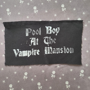 Może przedstawiać: Czarna prostokątna naszywka z białym napisem "Pool Boy At The Vampire Mansion" gotycką czcionką. Naszywka ma surową krawędź. Tło to wzorzysta tkanina z białymi różami i pająkami.