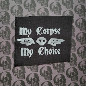 May include: A black fabric patch with the phrase "My Corpse My Choice" in white gothic lettering. The design includes a skull and wing graphic. The background features a repeating pattern of gray skulls on a purple background.