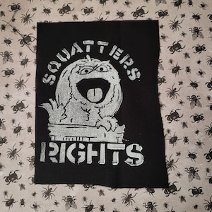Puede incluir: Parche de tela negra con las palabras "SQUATTERS RIGHTS" en negrita, blanco, con una fuente tipo plantilla. El parche presenta un personaje caricaturesco, parecido a un monstruo, con la boca muy abierta y la lengua fuera.