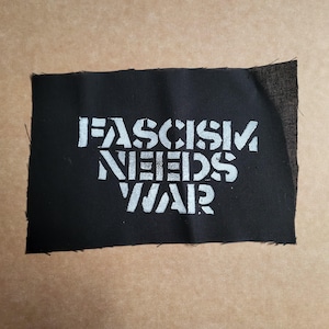 May include: A black fabric patch featuring the white text "FASCISM NEEDS WAR" in a bold, stencil font. The patch has raw edges and is displayed on a neutral background.