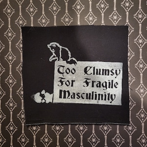 May include: Black fabric patch with white text that reads "Too Clumsy For Fragile Masculinity". A white cartoon bear is on top of the text.