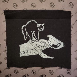 Puede incluir: Un parche bordado en blanco y negro que representa un gato negro de pie sobre una cámara de seguridad. El parche está sobre un fondo de tela negro.