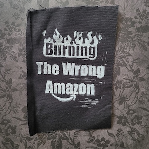 May include: Black patch with white text that reads "Burning The Wrong Amazon". The text is styled with flames above the words "Burning".