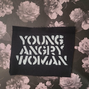 May include: A black fabric patch with the words "YOUNG ANGRY WOMAN" in stencil-style white lettering. The patch is set against a floral background with pink and white roses.