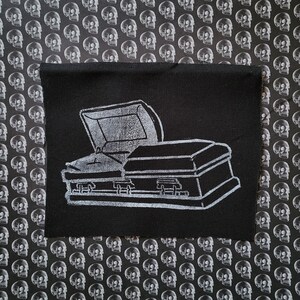 May include: Black rectangular patch featuring a white illustration of an open coffin. The coffin is depicted with a detailed outline, including the hinges and handles. The background is a repeating pattern of white skulls on a black surface.