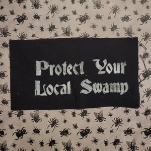 May include: A black patch with the text "Protect Your Local Swamp" in white, set against a patterned background of insects. The background features a repeating pattern of spiders and flies in black on a light brown surface.