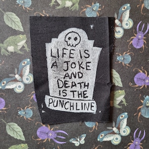 Puede incluir: Parche de tela negro con texto blanco que dice "Life is a joke and death is the punchline". El texto está en una lápida blanca con una calavera en la parte superior.