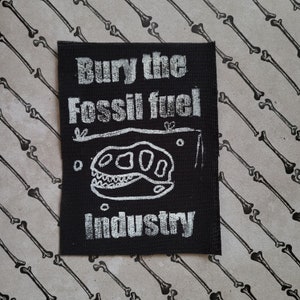 Puede incluir: Parche de tela negro con texto blanco que dice "Bury the Fossil Fuel Industry". Un cráneo de dinosaurio de dibujos animados blanco se encuentra en el centro del parche.