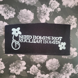 Może przedstawiać: Czarna prostokątna naszywka z napisem "SEED BOMBS NOT NUCLEAR BOMBS" w kolorze jasnoniebieskim. Naszywka posiada grafikę kwiatową po obu stronach tekstu oraz grafikę kwiatową z symbolem po lewej stronie.