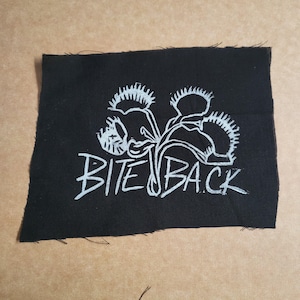 Può includere: Toppa in tessuto nero con un'illustrazione bianca di piante carnivore e la scritta "BITE BACK". Il disegno è in stile disegnato a mano. La toppa ha un bordo grezzo.