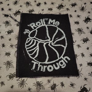 May include: Black fabric patch with white embroidered text that reads "Roll Me Through". The design features a stylised image of a wheel with spokes.