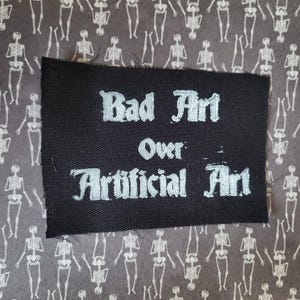May include: A black fabric patch with the words "Bad Art Over Artificial Art" in white, stylized lettering. The patch is set against a background of a repeating pattern of white skeleton figures on a gray background.