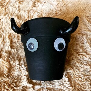 Può includere: Vaso di terracotta nero con occhietti e corna nere, che ricorda un toro o una mucca. Il vaso è su una superficie soffice di colore marrone chiaro.