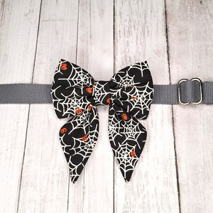 May include: A black bow tie with a white spiderweb and orange pumpkin pattern, attached to a grey adjustable collar with a silver D-ring.