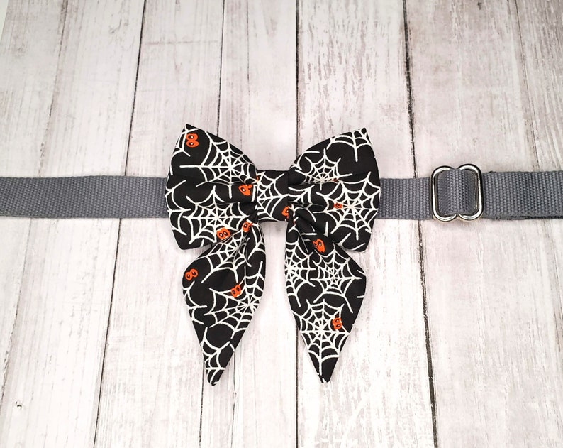 May include: A black bow tie with a white spiderweb and orange pumpkin pattern, attached to a grey adjustable collar with a silver D-ring.