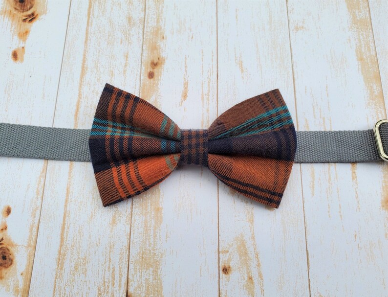 May include: A plaid bow tie with orange, blue, and black colors. The bow tie is attached to a gray strap.
