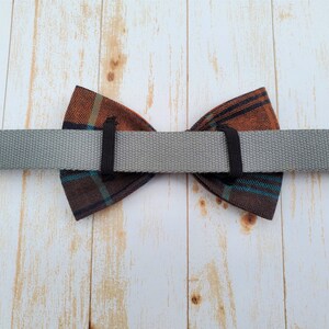 May include: A brown and blue plaid bow tie with a gray adjustable strap. The bow tie is attached to the strap with black elastic.