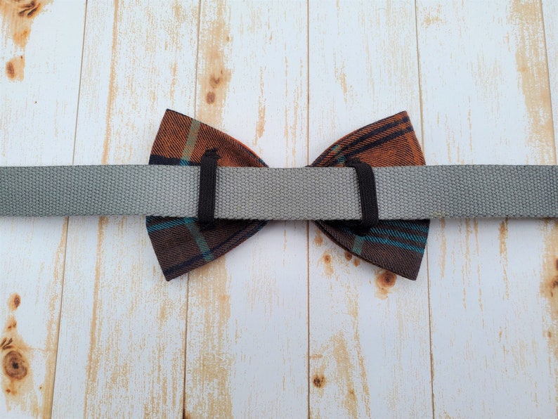 May include: A brown and blue plaid bow tie with a gray adjustable strap. The bow tie is attached to the strap with black elastic.