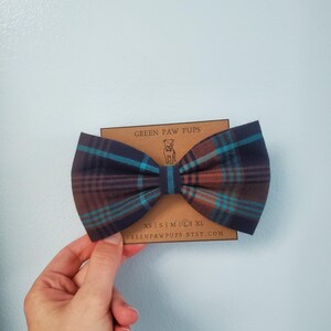 May include: A brown, blue, and orange plaid bow tie for a dog. The bow tie is on a brown card with the text "GREEN PAW PUPS" and "GREENPAWPUPS.ETSY.COM".